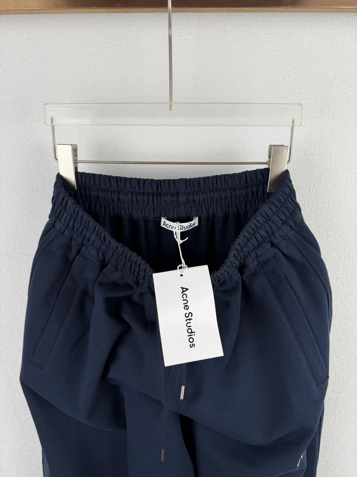 ACNE STUDIOS embroidered logo fleece sweatpants wide-leg trousers. Fabric: 32-count double yarn top + 16-count terry bottom yarn G The veil is made of 32-count fine yarn. The high-count yarn is smooth and delicate. After weaving, it forms a smooth and compact surface. It not only resists pilling and wear-resistant, but also gives the fabric a touch as delicate as skin. The base yarn is made of 16-count roving. The low-count yarn is fluffy and hairy. It naturally forms a rich terry layer during the weaving process, providing a sufficient source of fiber for subsequent sanding. This combination of fine surfaces and thick soles strikes a perfect balance between warmth and skin-friendliness. Mechanical sanding is the soul process of this fabric. By rubbing the bottom surface of the roller wrapped with emery leather at high speed, the fiber ends of the 16-count roving are evenly pulled out and cut off, forming a dense and uniform short pile layer. The smoothness and neatness of needle embroidery and the soft velvet feel of brushed fabrics form a material dialogue between hardness and softness. The embroidery area is smooth and crisp, with sharp lines, like delicate inscriptions carved on a rich suede canvas; while the surrounding brushed fabric presents a soft matte natural texture. The spacing between stitches is consistent and the strength is balanced to ensure that each stitch is placed accurately. This rigorous stitching structure makes the edges of the logo present a sharp and clear outline, and the strokes of the text are full and smooth. Even the curved turning points of the floral fonts can achieve a smooth transition through the connection of tight stitches.